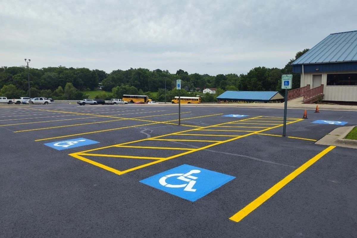 Freshly resurfaced lot with yellow stall lines, hatched access aisles and blue accessible bays, school buses parked beyond